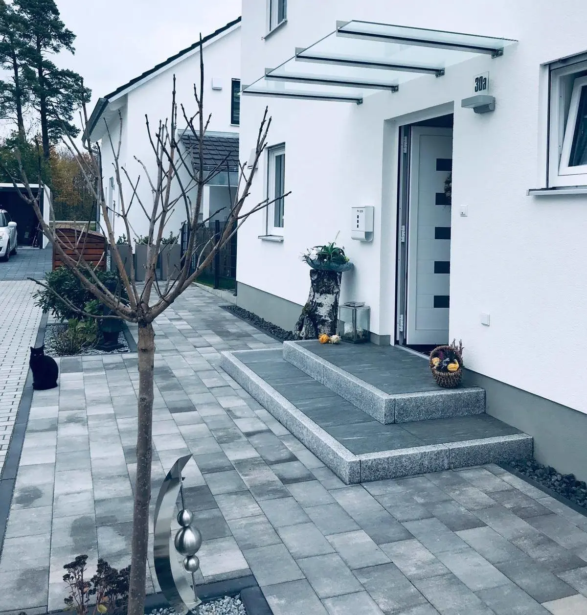 Ein modernes weißes Haus mit einer überdachten Veranda aus Glas und einem grau gefliesten Gehweg. Auf den Stufen steht eine Topfpflanze, ein blattloser Baum und am Weg sitzt eine schwarze Katze. In der Nähe des Eingangs steht eine Metallskulptur.