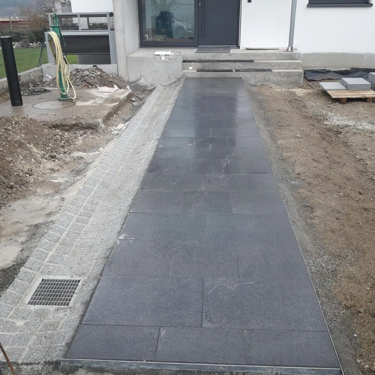 Ein neu angelegter Steinweg führt zum Eingang eines Hauses. Auf der einen Seite ist Kies und auf der anderen Seite Erde. Am Rand ist ein Abflussgitter zu sehen, im Hintergrund sind Baumaterialien zu sehen.