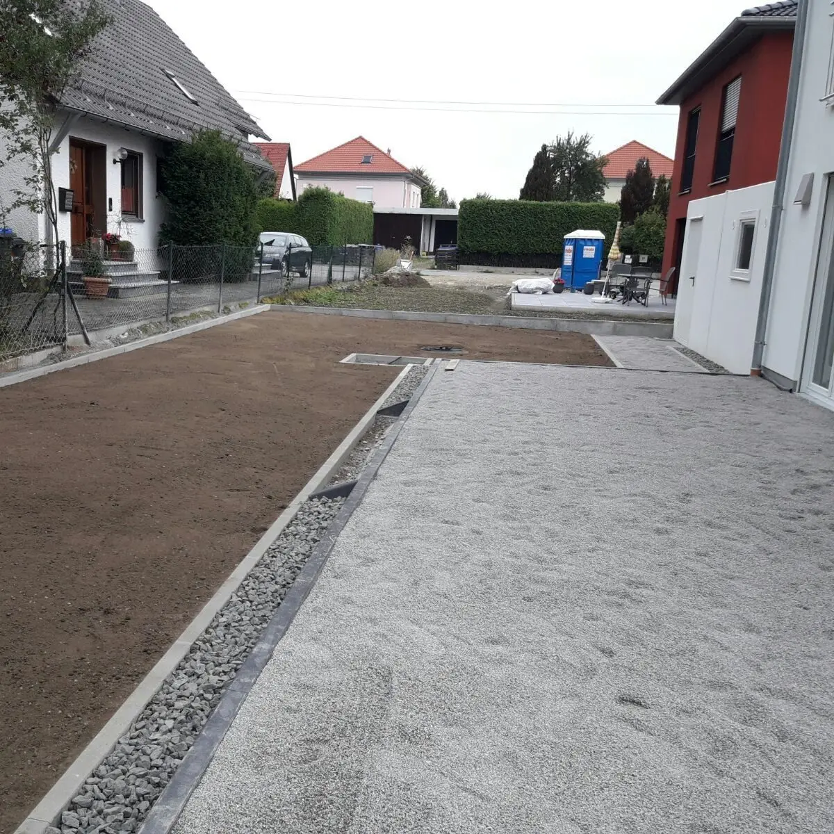 Ein Wohngebiet mit zwei Häusern, die durch eine Einfahrt getrennt sind. Der Hof auf der linken Seite ist mit Erde vorbereitet und die rechte Seite ist mit Kies bedeckt. Im Hintergrund sind eine tragbare Toilette und Baumaterialien zu sehen.