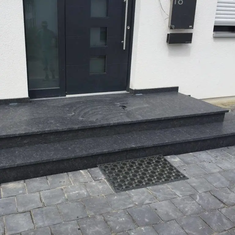 Schwarze Steinstufen führen zu einer modernen schwarzen Tür mit vier quadratischen Fenstern. Auf den Stufen liegt eine Fußmatte, und in der Tür ist schwach das Spiegelbild eines Mannes zu erkennen. Die Stufen sind von grauen Pflastersteinen umgeben.