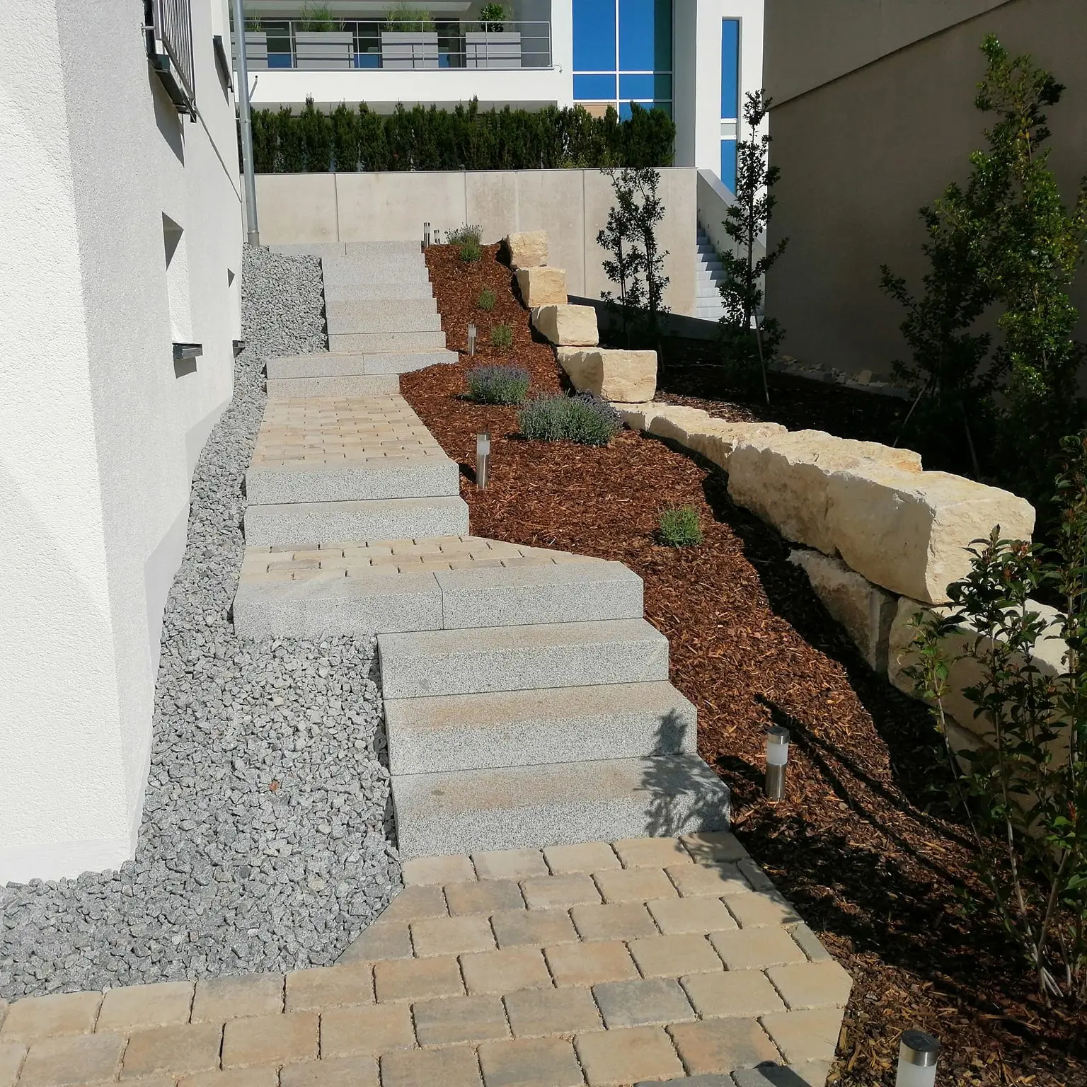 Ein angelegter Garten mit Steinstufen, umgeben von Mulch und Ziersteinen. Auf beiden Seiten sind kleine Pflanzen angeordnet, und Stützmauern aus Stein begrenzen den Bereich. Die Anlage grenzt an ein modernes weißes Gebäude.