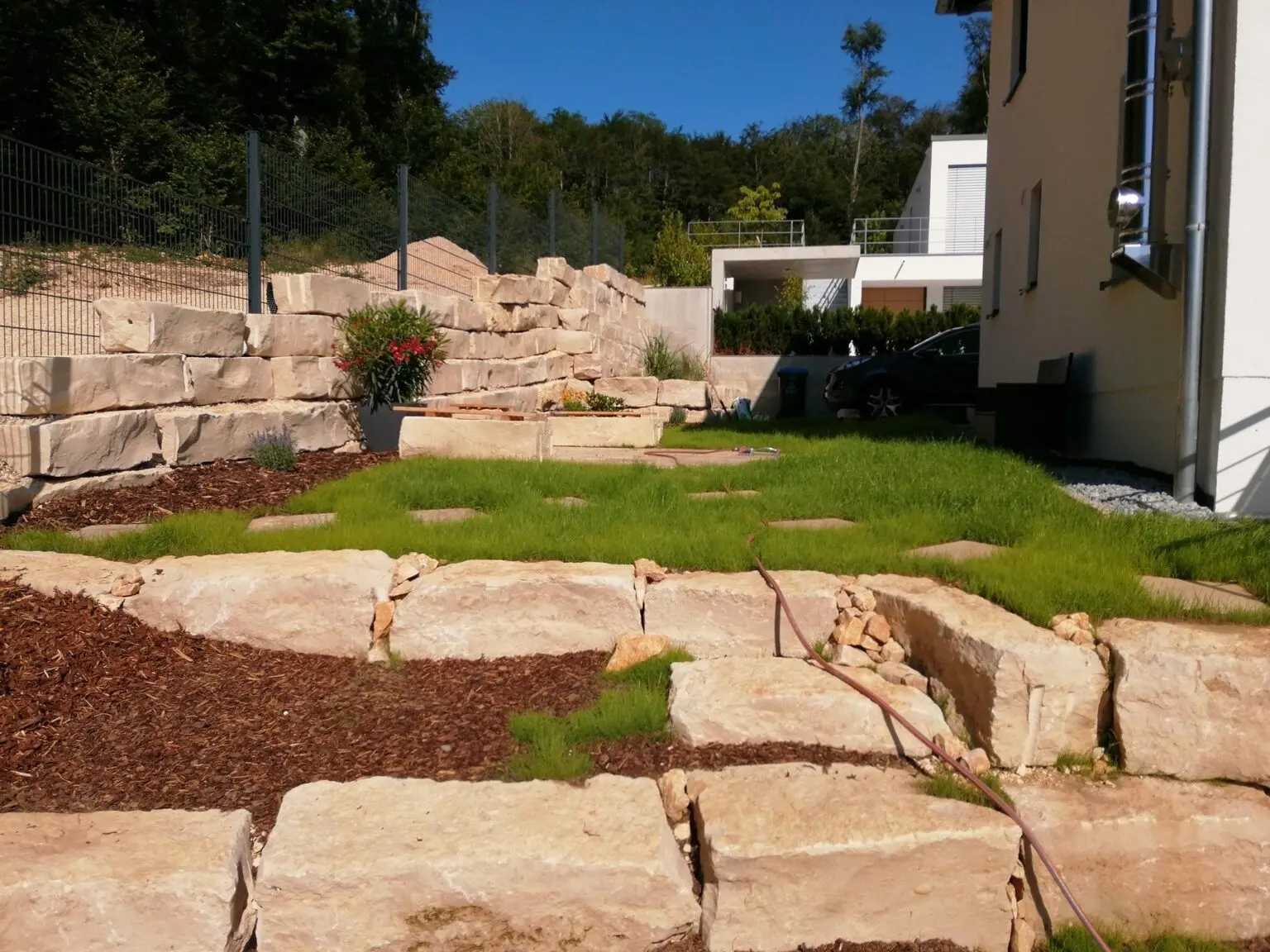 Ein angelegter Hinterhof mit gestapelten Steinmauern und gepflegten Rasenflächen. Mehrere Blumentöpfe stehen auf den Steinsimsen. Ein Gartenschlauch ist über den Rasen gelegt. Im Hintergrund sind ein Zaun und ein Haus zu sehen.