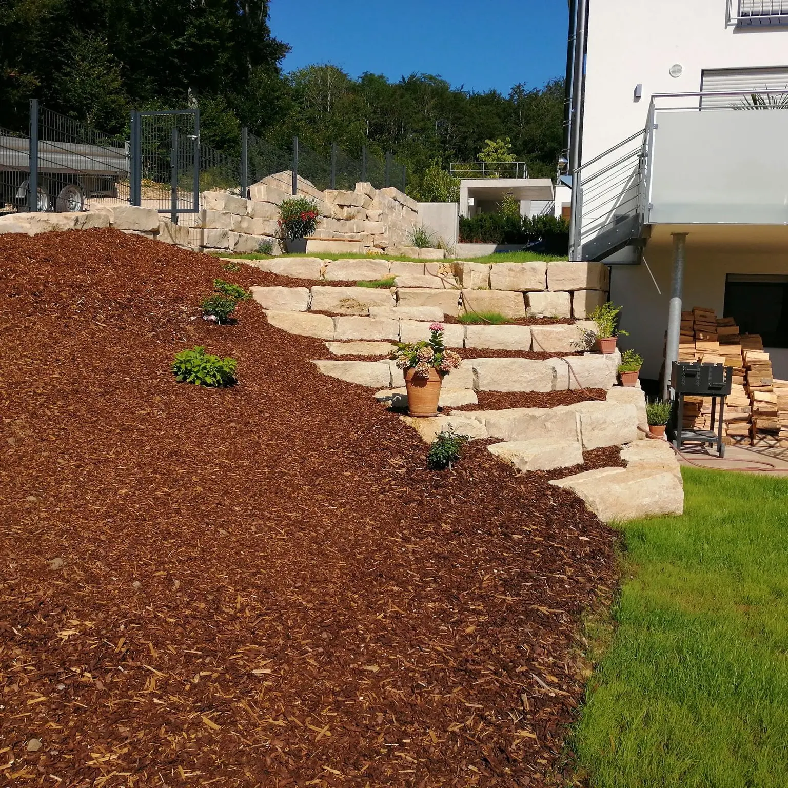 Ein abgestufter Garten mit großen Steinstufen, umgeben von Mulch und verschiedenen Grünpflanzen. Auf einer Stufe steht ein Topf mit Blumen. Rechts ist ein weißes Gebäude mit gestapeltem Brennholz unter einem Balkon zu sehen. Im Hintergrund sind ein Zaun und Bäume zu sehen.