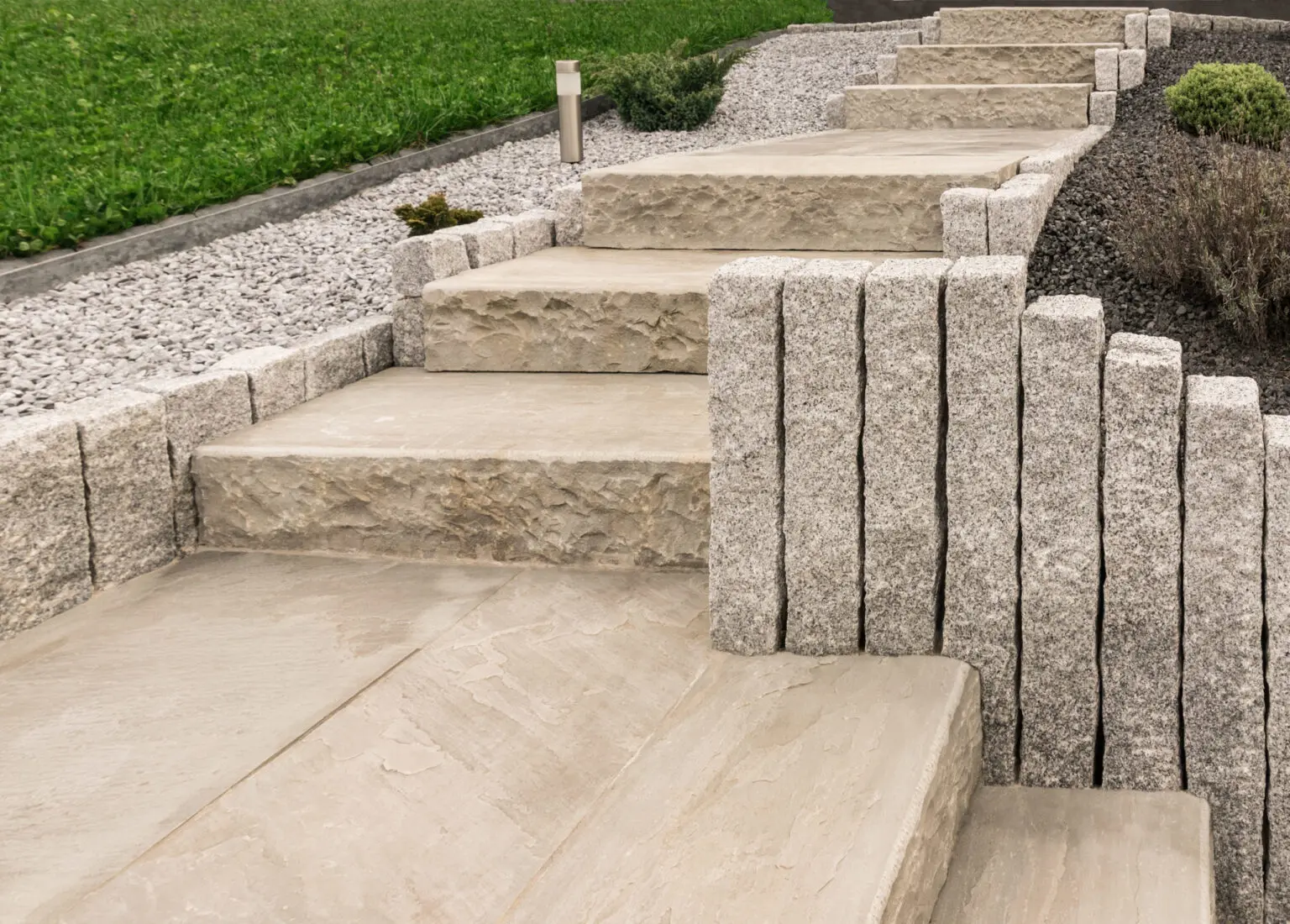 Steinstufen führen bergauf, gesäumt von vertikalen Steinplatten und mit Kies und Grünflächen bepflanzt. Der Weg steigt durch ordentlich angeordnete Gartenelemente an und erzeugt ein strukturiertes und aufgeräumtes Erscheinungsbild.