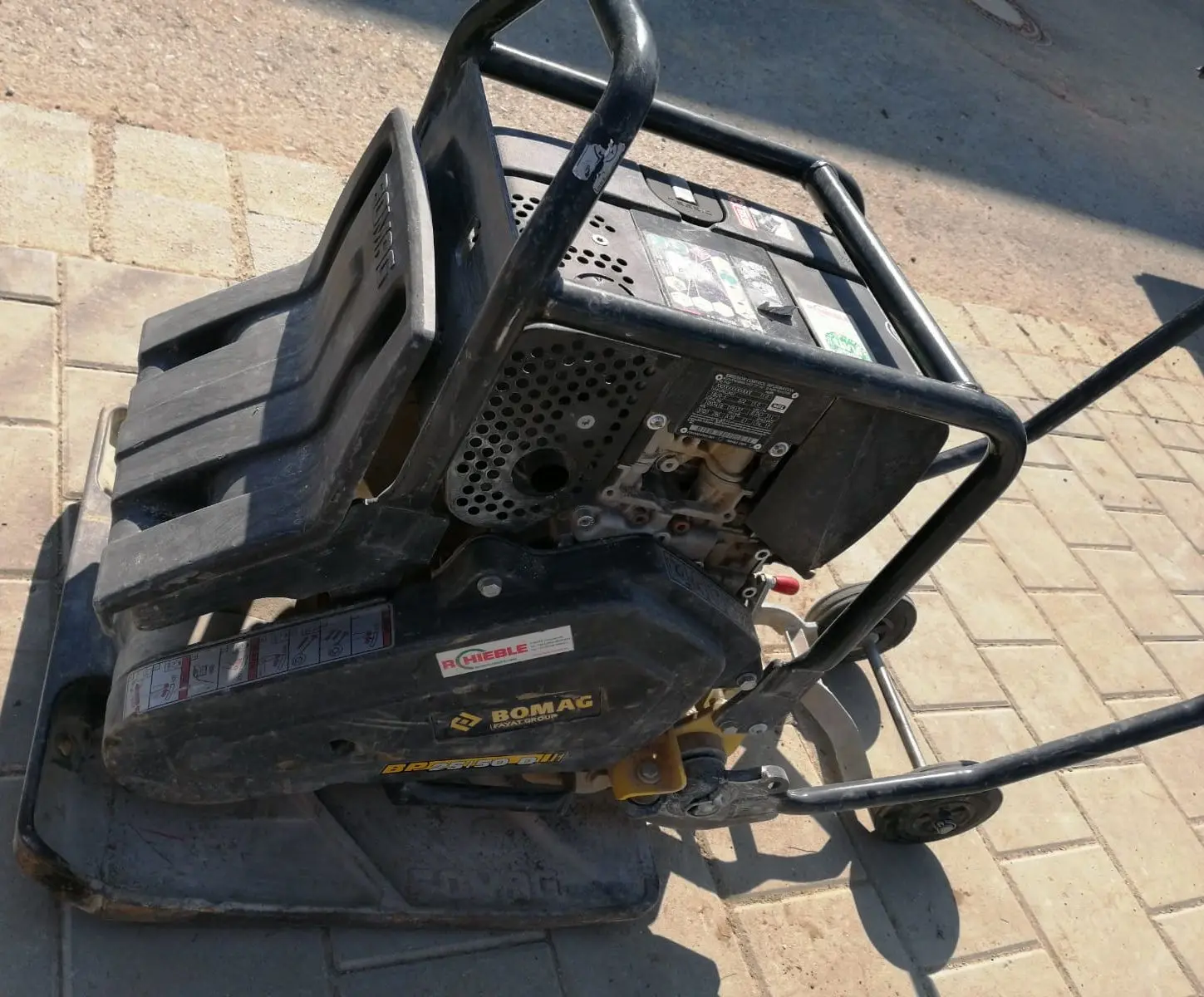 Bild einer Verdichtungsmaschine von BOMAG auf einer gepflasterten Fläche. Die Maschine hat einen schwarzen Metallrahmen mit Griffen und sichtbarem Markenzeichen. Sie wird zum Verdichten von Erde, Asphalt und anderen Materialien bei Bauarbeiten verwendet.