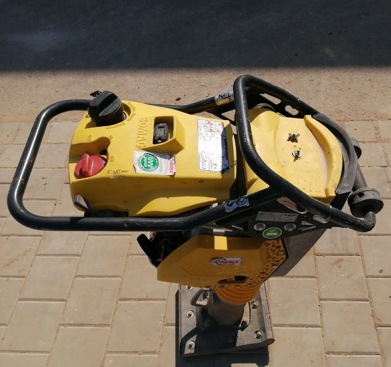 Eine Draufsicht auf eine gelbe Verdichtungsmaschine auf einer gepflasterten Fläche. Die Maschine hat einen runden Tankdeckel, Bedienhebel und verschiedene Etiketten und Aufkleber. Der Boden darunter besteht aus hellbraunen rechteckigen Ziegeln.