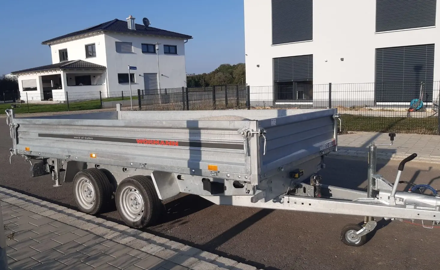Ein zweiachsiger Metallanhänger ist auf einer gepflasterten Stadtstraße geparkt. Er steht in der Nähe von zwei modernen, weißen Gebäuden mit großen Fenstern und einem Hofbereich. Der Anhänger scheint zum Transport von Waren oder Materialien zu dienen.