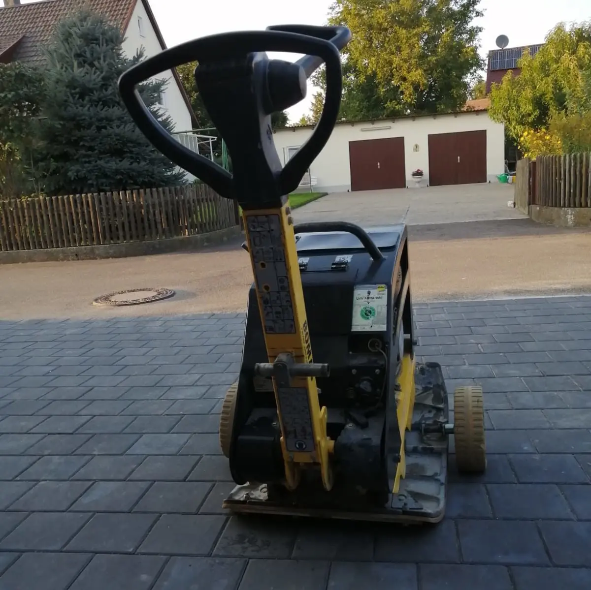Eine Nahaufnahme eines kompakten Bodenverdichters auf einer gepflasterten Einfahrt. Die Maschine verfügt über einen Griff zur Bedienung und weist ein robustes Design auf, das für Bau- oder Landschaftsbauarbeiten geeignet ist. Im Hintergrund sind Wohnhäuser und Bäume zu sehen.