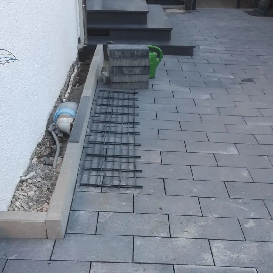 Gezeigt wird eine gepflasterte Fläche mit grauen, rechteckigen Fliesen. Links ist ein schmaler Pflanzkübel oder Graben mit freiliegenden Rohren zu sehen, der teilweise von einem Metallgitter bedeckt ist. In der Nähe befinden sich ein Stapel Betonblöcke und eine grüne Gießkanne. Stufen führen auf eine höhere Ebene.