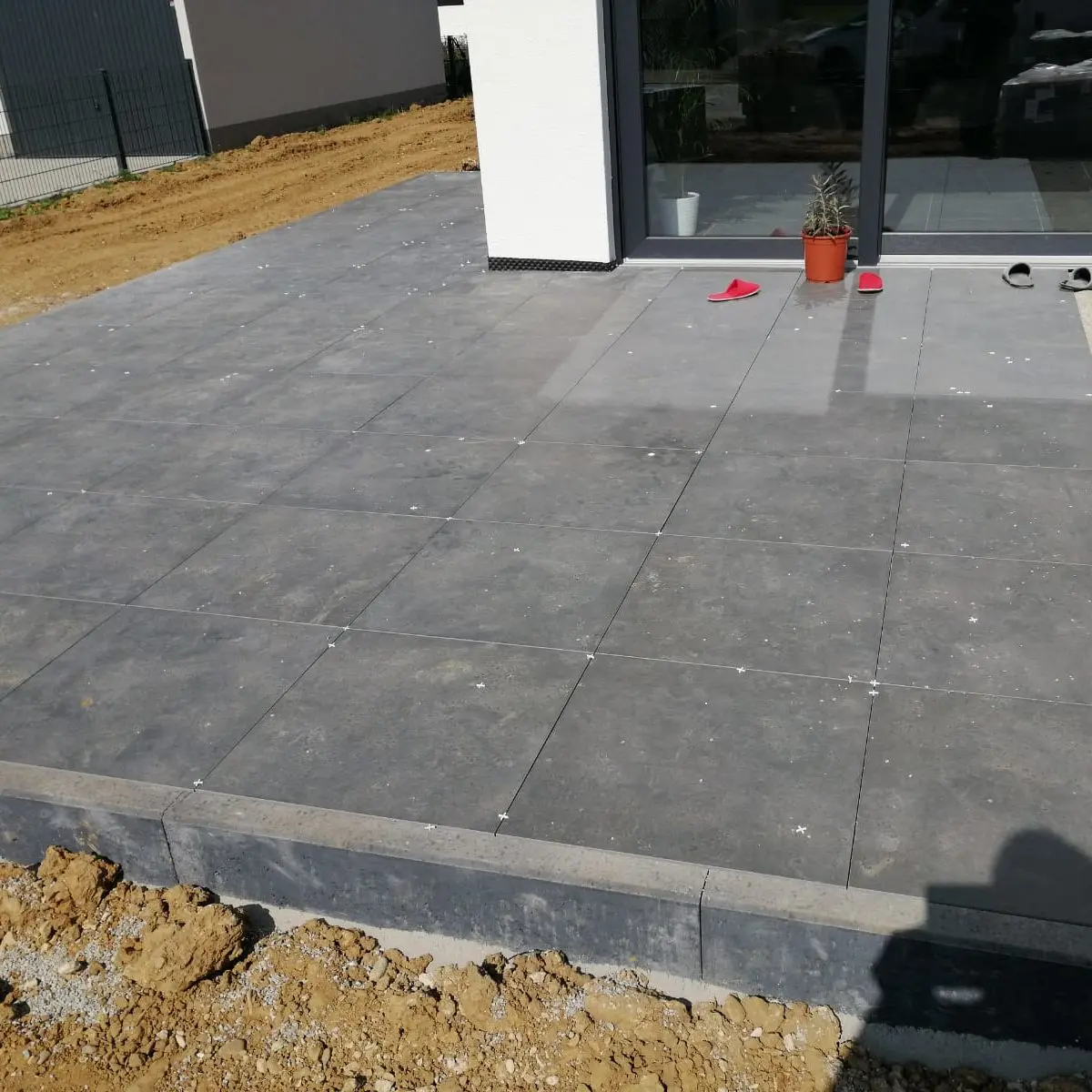 Von einem Gebäude aus erstreckt sich eine Terrasse mit großen grauen Fliesen. Ein Paar rote Sandalen und eine Pflanze in einem Topf stehen neben den Glasschiebetüren. Der Boden um die Terrasse herum ist kahl und im Vordergrund ist ein Schatten zu sehen.