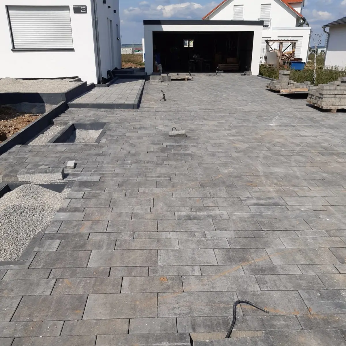 Eine im Bau befindliche Auffahrt mit grauen Pflastersteinen. Im Vordergrund sind teilweise verlegte Steine zu sehen, in einigen Bereichen mit losem Kies. Im Hintergrund sind eine Garage und ein weißes Gebäude zu sehen. Auf der rechten Seite sind Stapel mit Baumaterialien zu sehen.
