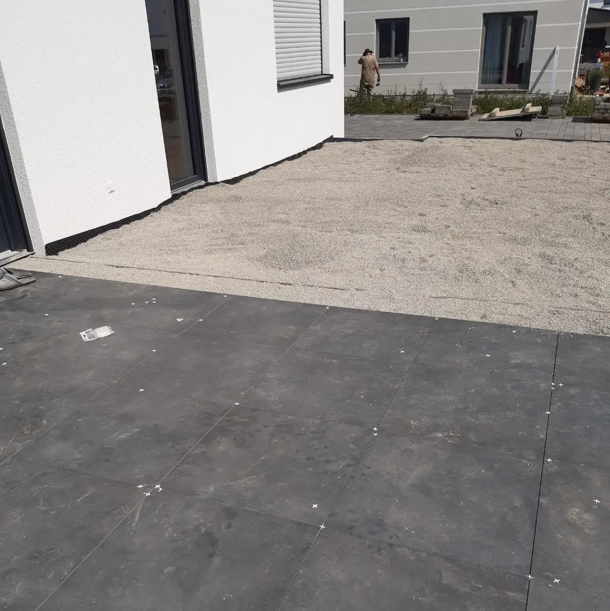 Ein Terrassenbereich wird gerade umgebaut. Im Vordergrund sind frisch verlegte dunkle Steinfliesen zu sehen, während im Hintergrund ein Abschnitt mit Kies zu sehen ist. Eine teilweise sichtbare Person arbeitet in der Nähe eines modernen Gebäudes. Werkzeuge und Materialien liegen verstreut herum.