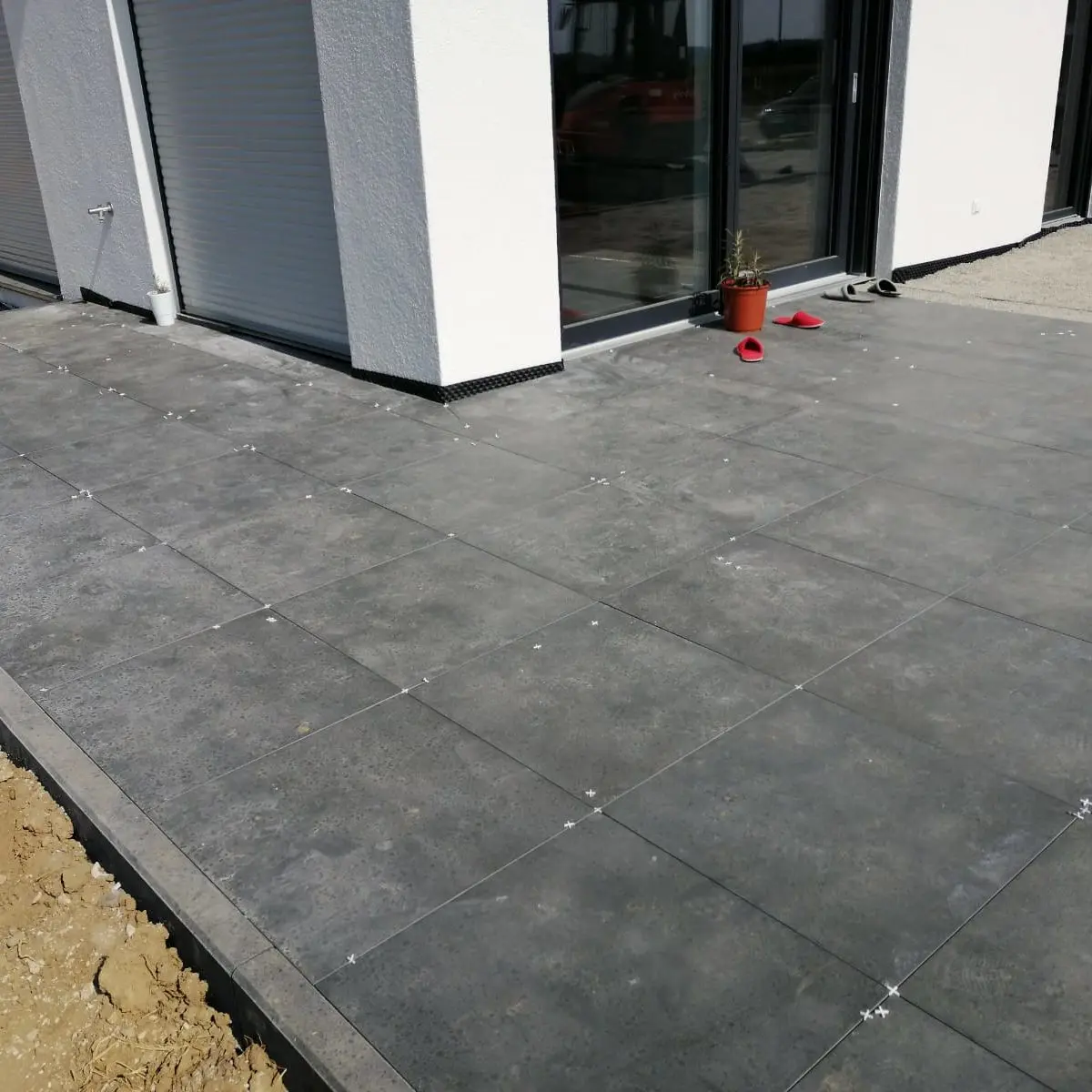 Gezeigt wird ein modernes Gebäude mit großen Fenstern. Der Bereich verfügt über eine Außenterrasse mit grauen quadratischen Fliesen. In der Nähe des Eingangs stehen ein kleiner roter Blumentopf und rote Schuhe. Der Boden neben den Fliesen ist nackte Erde.