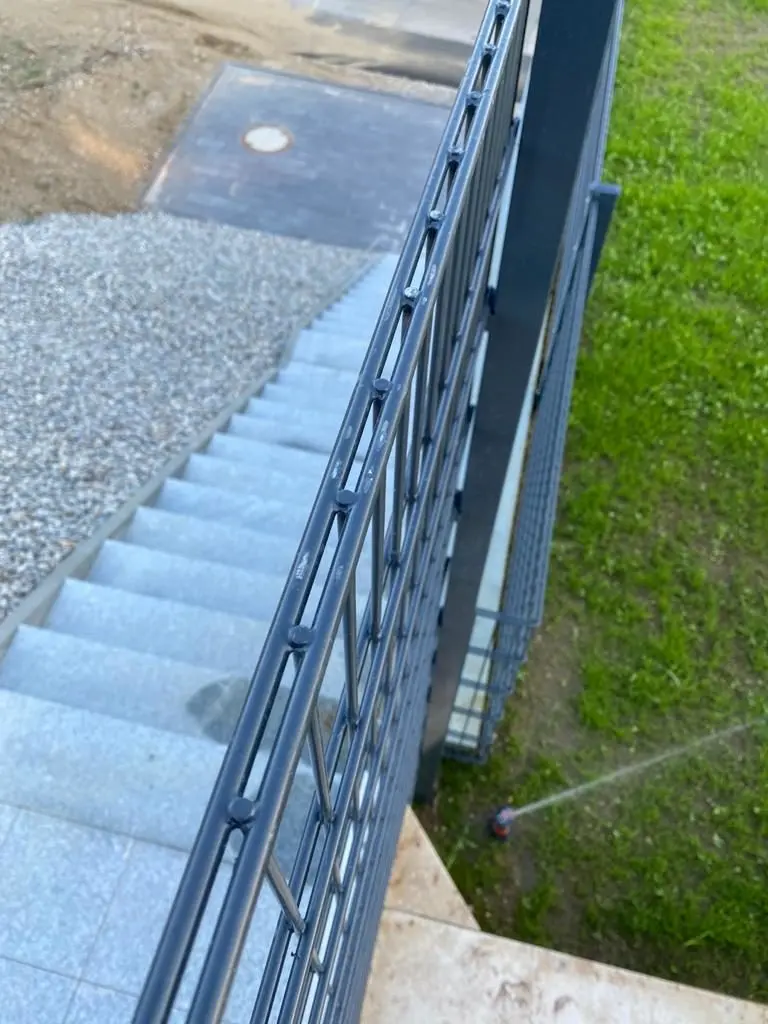 Blick von oben auf eine Außentreppe mit einem Metallgeländer auf der linken Seite. Die Treppe führt hinunter zu einer Kiesfläche. Auf der rechten Seite ist eine Rasenfläche mit einem Sprinkler zu sehen, der Wasser versprüht.