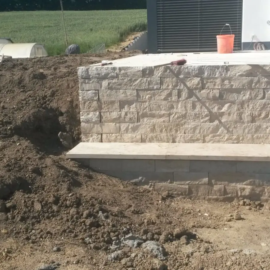 Bauszene mit einer Steinmauer, vor der Erde aufgehäuft ist. Von der Mauer geht eine flache Fläche ab, möglicherweise ein Sitzbereich. Auf der Mauer stehen ein roter Eimer und ein Werkzeug. Im Hintergrund sind ein Feld und ein Teil eines Gebäudes zu sehen.