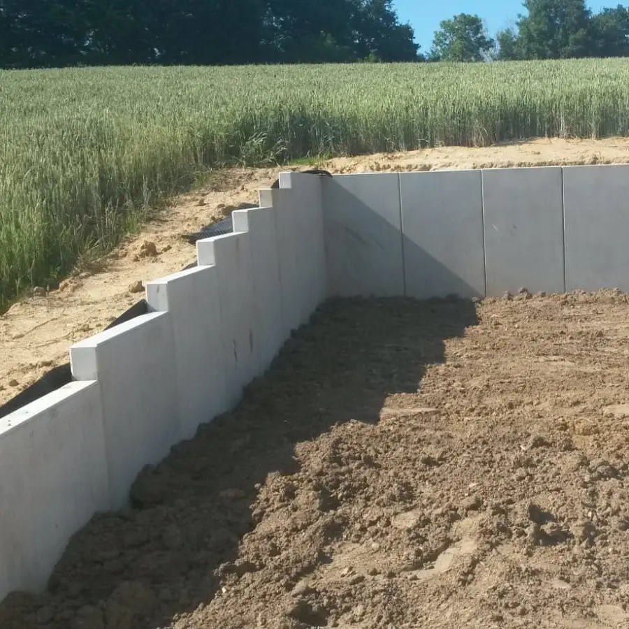 Betonstützmauer im Bau mit Erdauffüllung. Stufen an der Mauer führen hinauf zu einer Wiese. Im Hintergrund sind klarer blauer Himmel und Bäume zu sehen.