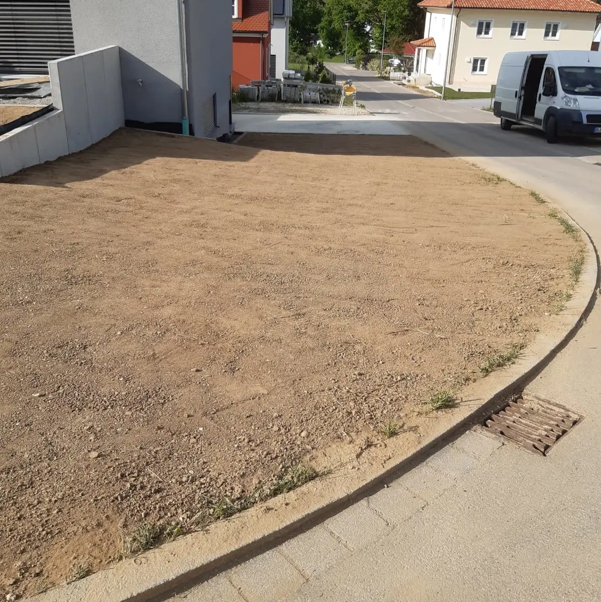 An einer Straßenecke eines Wohnhauses liegt ein mit Erde bedeckter Vorgarten neben einer asphaltierten Straße. Im Hintergrund sind mehrere Häuser zu sehen. Auf der rechten Straßenseite parkt ein weißer Lieferwagen. In der Nähe des Bordsteins befindet sich ein Abwasserkanal.