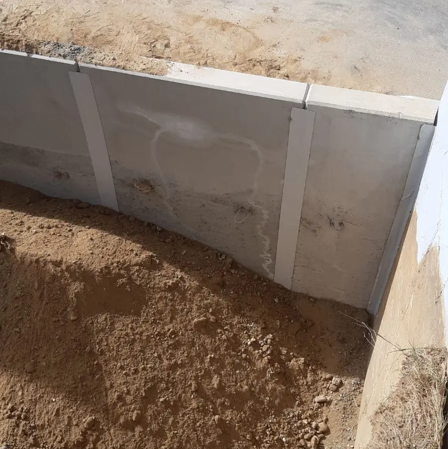 Eine Stützmauer aus Beton ist zu sehen, hinter der der Boden sichtbar ist. Der Boden ist uneben und mit Flecken aus losem Schmutz und kleinen Steinen übersät. Die Oberseite der Mauer ist glatt und weist mehrere Markierungen und Löcher auf ihrer Oberfläche auf.