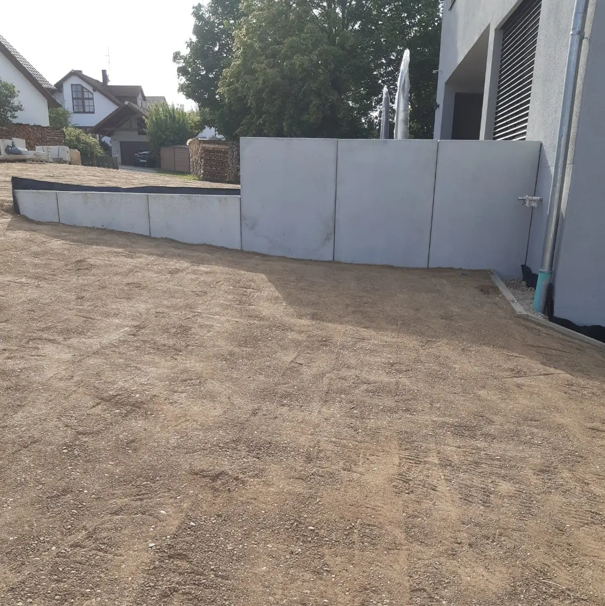Eine Kiesauffahrt führt zu einem hellgrauen Gebäude mit modernem Design. An der Seite des Gebäudes verläuft eine Betonmauer. Im Hintergrund sind ein Holzzaun und einige Bäume sowie ein Nachbarhaus zu sehen.