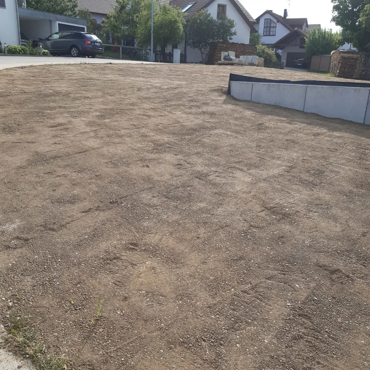 Ein unbefestigtes Grundstück in einem Wohngebiet mit ein paar Häusern und einem geparkten Auto im Hintergrund. Der Boden ist größtenteils kahl, mit einer kleinen erhöhten Betonstruktur auf der rechten Seite. In der Nähe der Gebäude sind Bäume und Sträucher sichtbar.