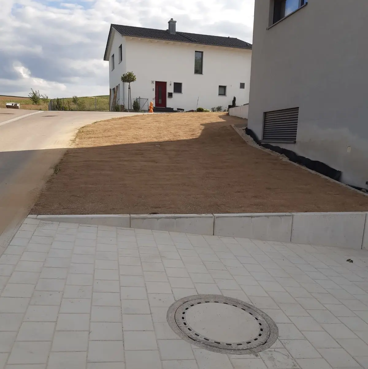 Ein neu errichtetes Haus mit weißer Fassade und roter Tür steht neben einer Straße. Der angrenzende Hof ist mit getrockneter Erde bedeckt, ohne sichtbare Vegetation. Ein weiteres Gebäude mit einer grauen Wand ist auf der rechten Seite teilweise sichtbar. Der Himmel scheint teilweise bewölkt zu sein.