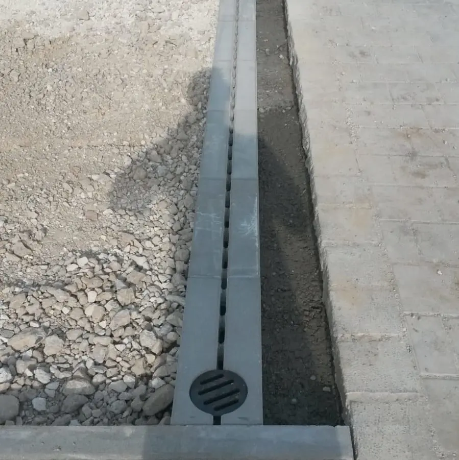Ein Beton-Abflusskanal mit Schlitzabdeckung verläuft rechts neben einer gepflasterten Fläche. Auf der linken Seite sind ein Kieshaufen und blanker Boden zu sehen. Im Vordergrund ist ein kreisrunder Abfluss zu sehen.