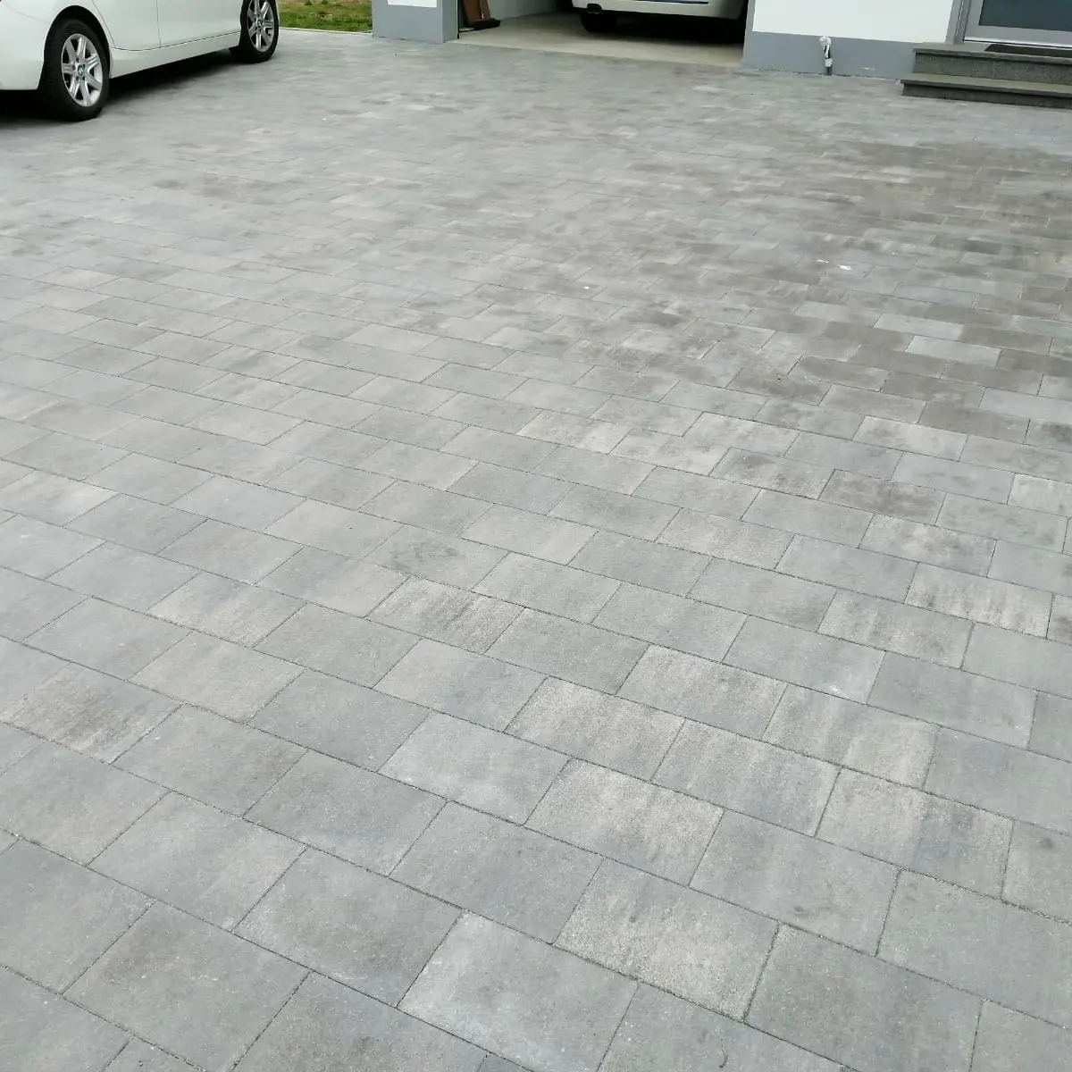 Eine Auffahrt besteht aus gleichmäßig verlegten grauen Pflastersteinen. Zwei weiße Autos sind zu sehen; eines ist teilweise zu sehen und ein anderes steht in einer Garage. Der Bereich ist sauber und gut gepflegt.