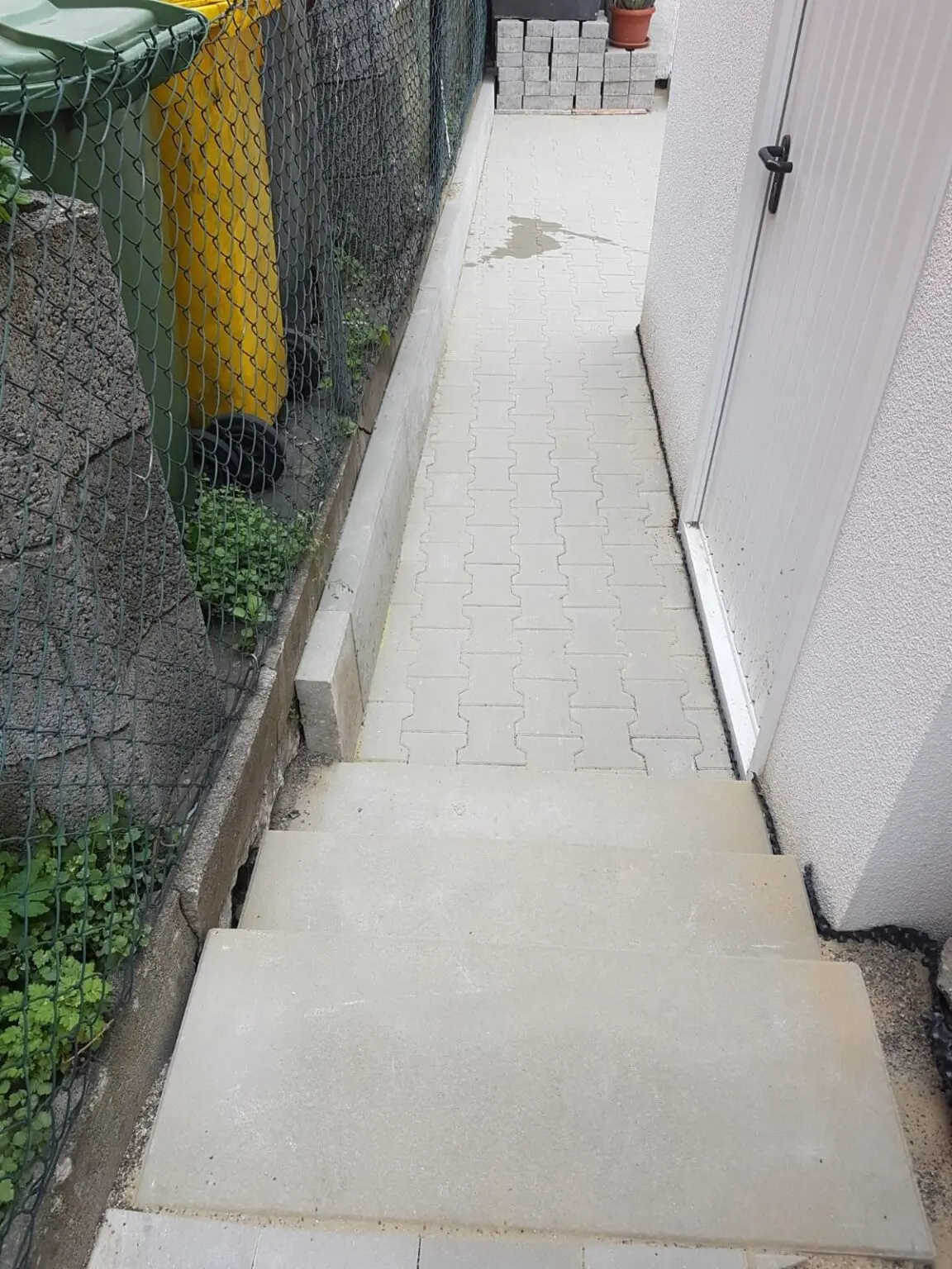 Ein schmaler Betonweg mit Stufen führt zu einer geschlossenen weißen Tür auf der rechten Seite. Ein grüner Maschendrahtzaun grenzt an die linke Seite, neben grünen Pflanzen. In der Nähe des Zauns sind ein gelber und ein grüner Mülleimer zu sehen. Im Hintergrund sind Stapel weißer Blöcke zu sehen.