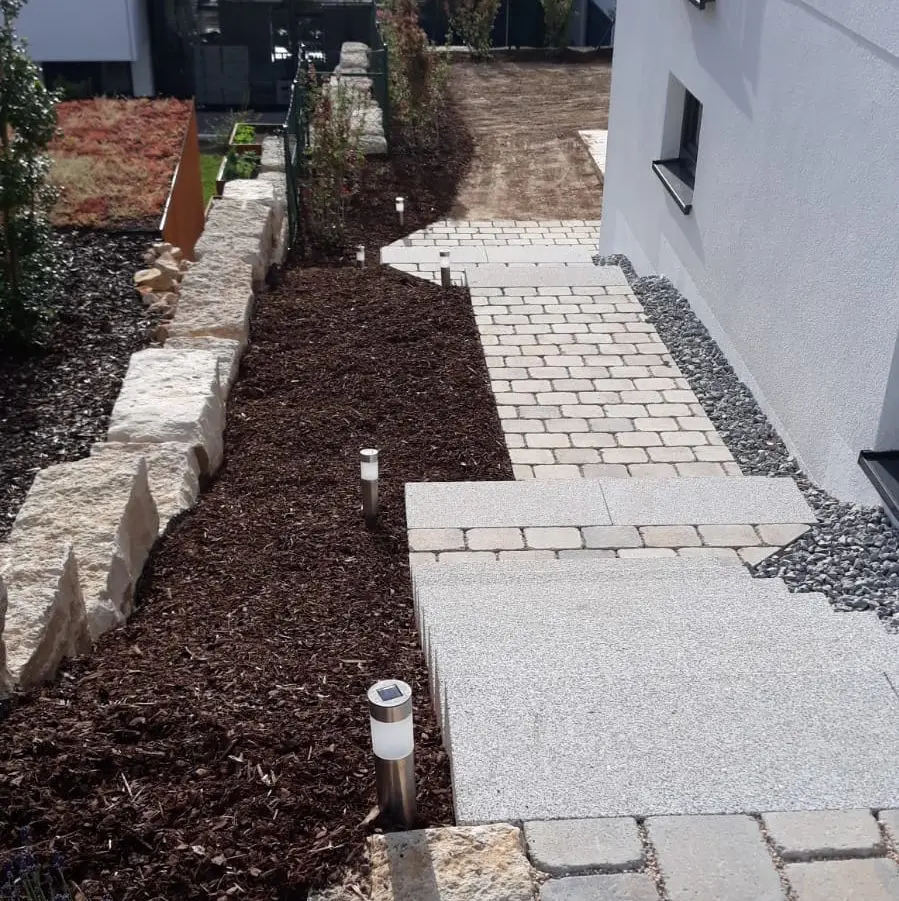 Ein mit Steinfliesen ausgelegter und von niedrigen Steinmauern gesäumter Gartenweg. Auf einer Seite befinden sich Mulch und kleine Sträucher, während solarbetriebene Lampen gleichmäßig entlang des Weges verteilt sind. Auf der rechten Seite ist ein Gebäude mit weißen Wänden zu sehen.