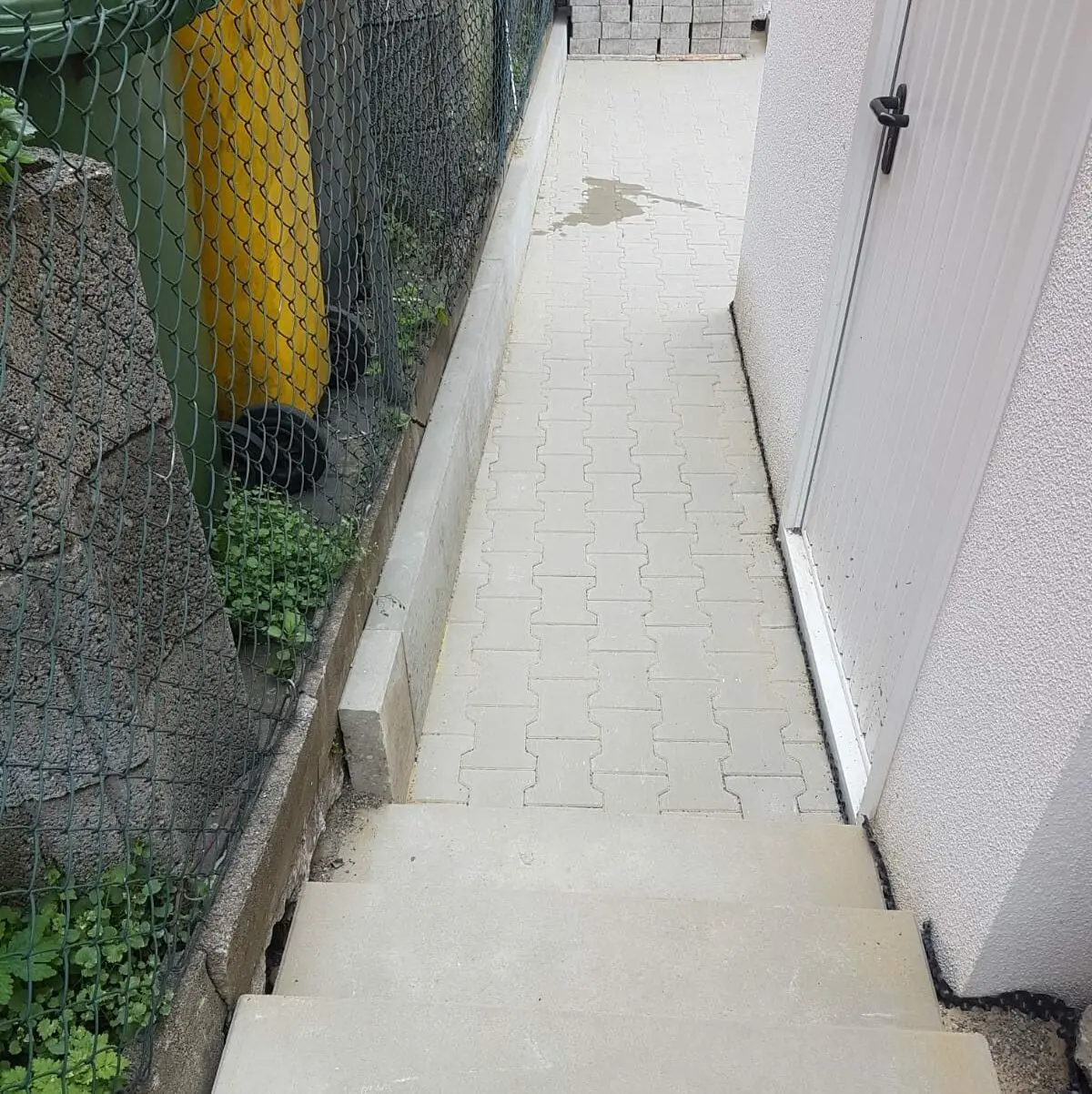 Ein schmaler Weg mit Verbundpflaster führt zu einer geschlossenen weißen Tür. Links verläuft parallel zum Weg ein grüner Maschendrahtzaun, und eine Reihe von Mülltonnen ist zu sehen. Vom Rand des Weges führen Stufen hinunter.