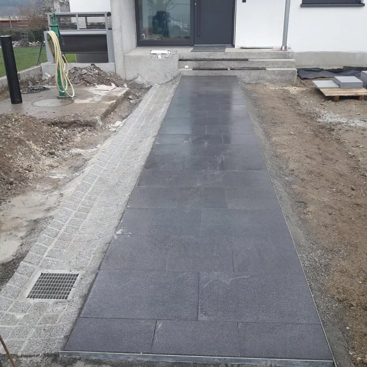 Ein neu angelegter Steinweg führt zum Eingang eines Hauses. Auf der einen Seite ist Kies und auf der anderen Seite Erde. Am Rand ist ein Abflussgitter zu sehen, im Hintergrund sind Baumaterialien zu sehen.