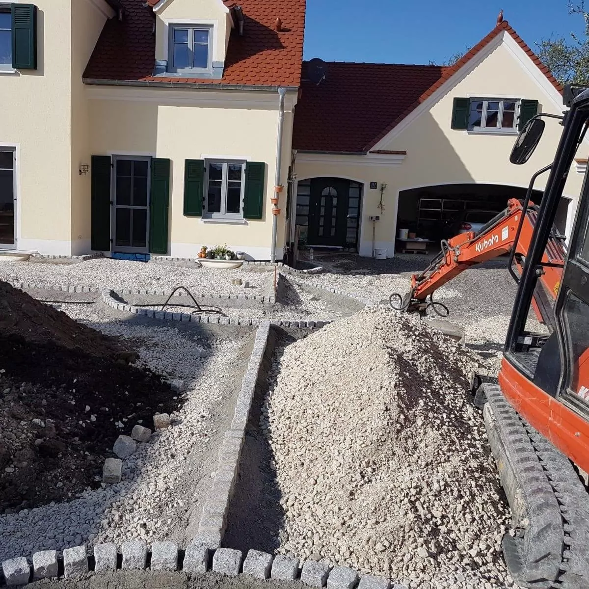 Eine Baustelle vor einem Haus mit rotem Dach, auf der ein kleiner Bagger Kies bewegt. Die Fläche ist mit Steinrändern umrandet, einige Fenster des Hauses haben grüne Fensterläden. Der Boden ist größtenteils mit Kies und Erde bedeckt.