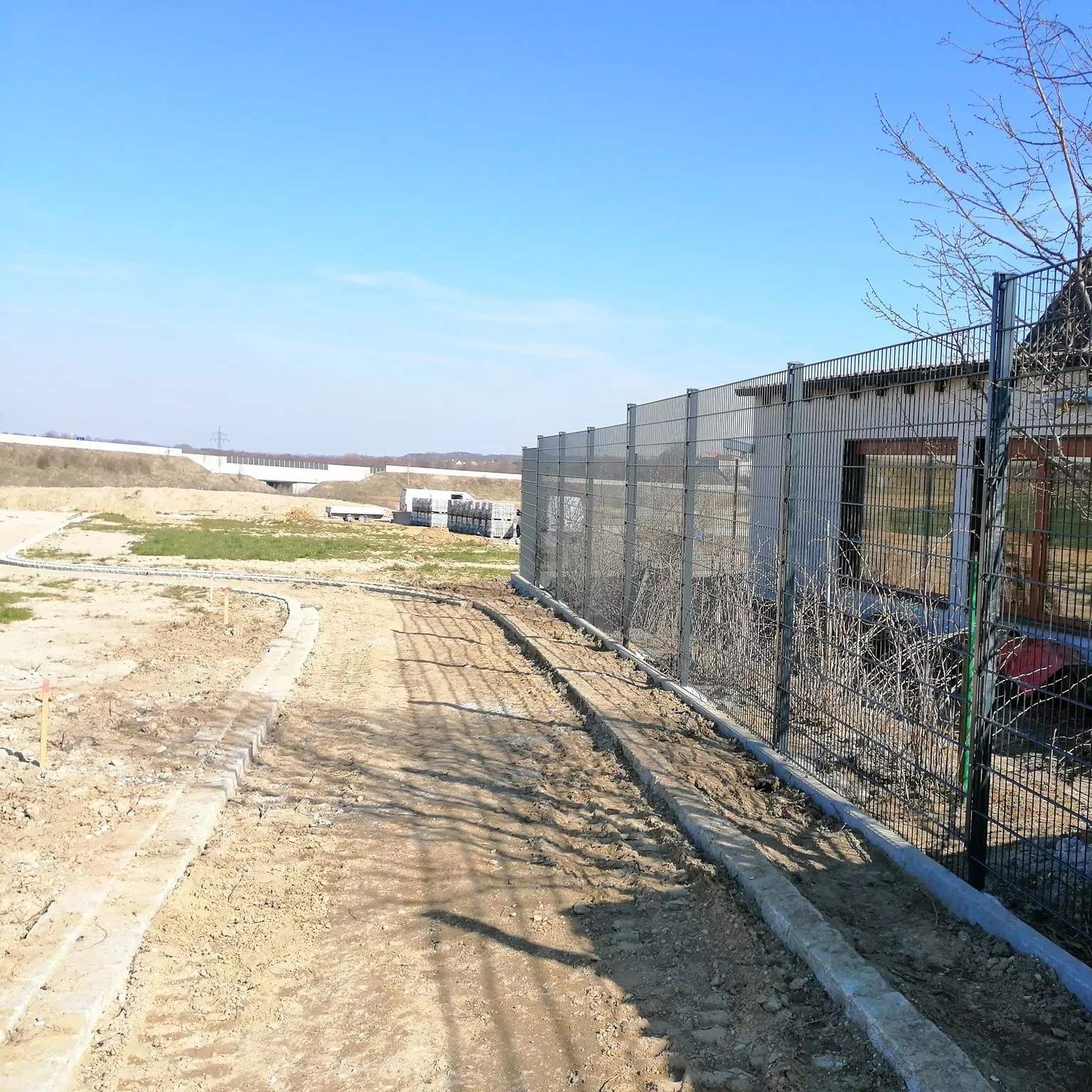 Unter einem klaren, blauen Himmel schlängelt sich eine unbefestigte Straße an einem Metallzaun vorbei. Der Zaun grenzt an eine Baustelle mit verstreuten Materialien. Die umliegende Landschaft ist offen, mit teilweise kahlem Boden und spärlicher Vegetation.
