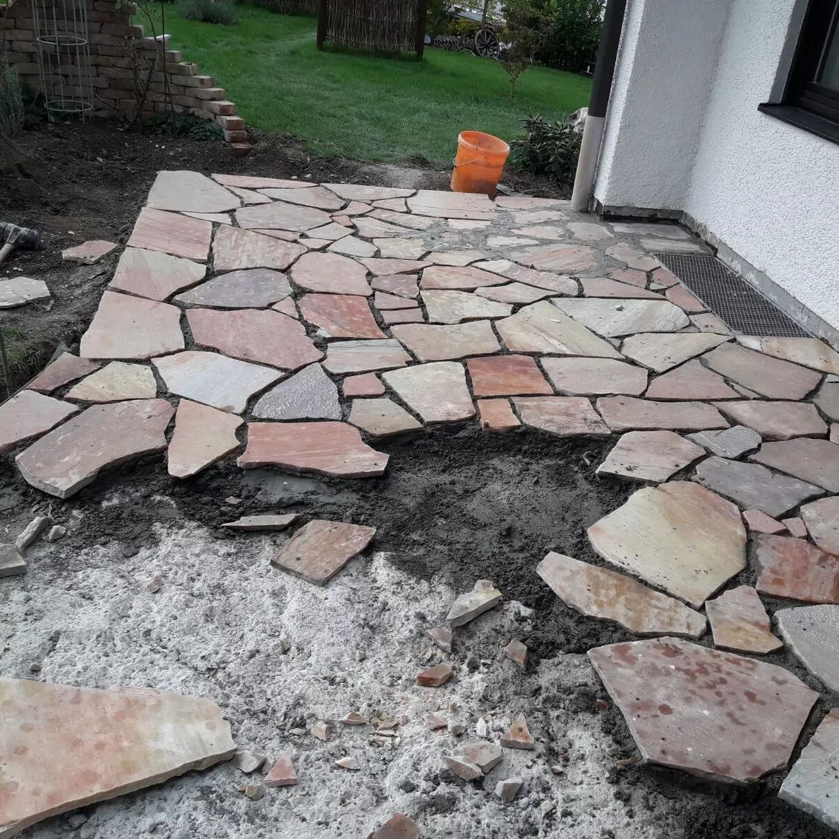 Gezeigt wird eine teilweise fertiggestellte Terrasse aus unregelmäßig geformten Steinplatten. Einige der Steine liegen noch an ihrem Platz, andere sind in der Nähe aufgestapelt. Im Hintergrund sind ein orangefarbener Eimer und Baumaterialien zu sehen.