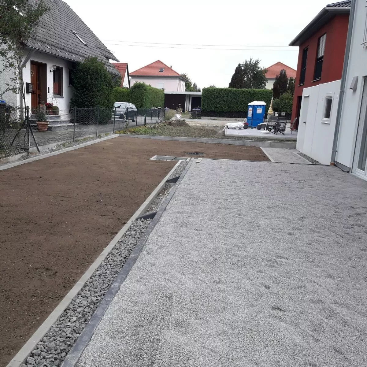 Ein Wohngebiet mit zwei Häusern, die durch eine Einfahrt getrennt sind. Der Hof auf der linken Seite ist mit Erde vorbereitet und die rechte Seite ist mit Kies bedeckt. Im Hintergrund sind eine tragbare Toilette und Baumaterialien zu sehen.