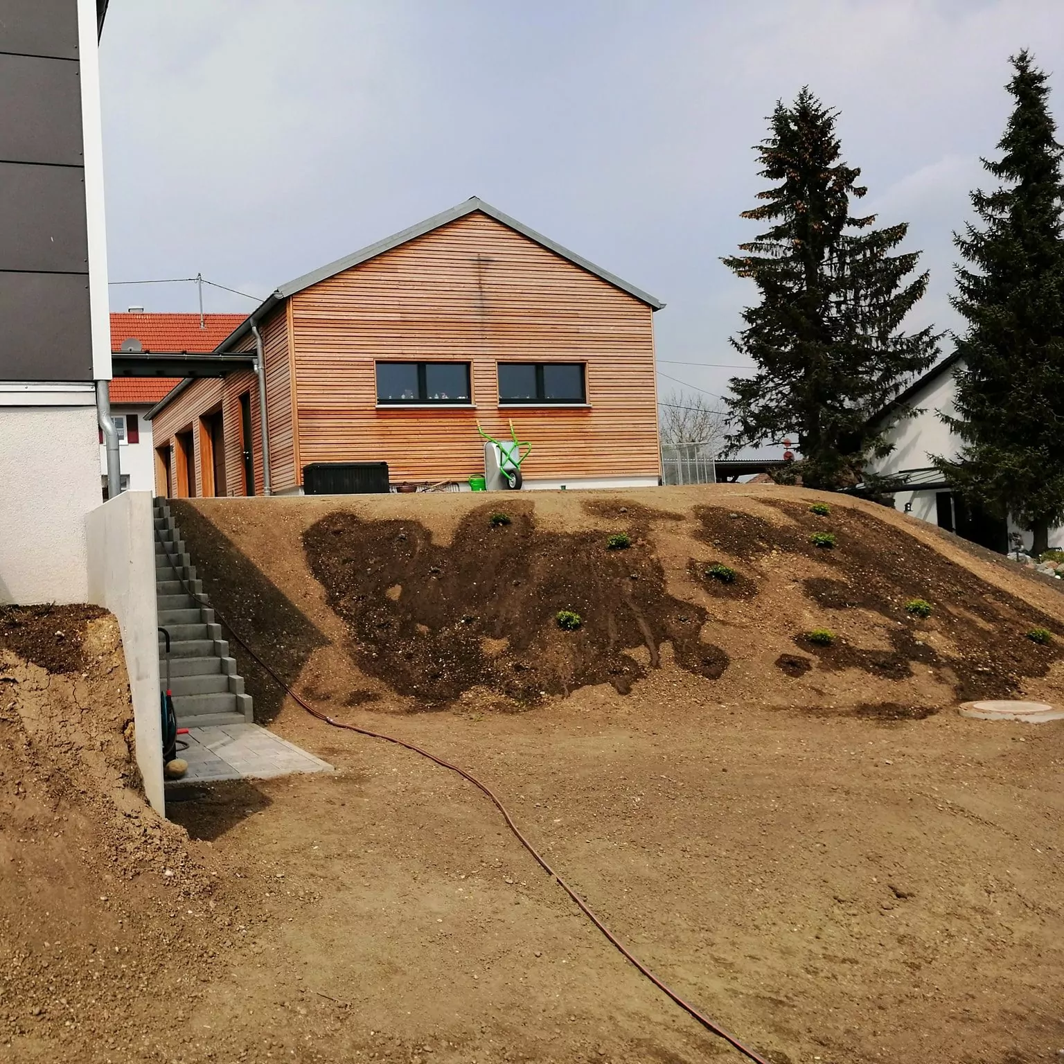 Ein Haus mit Holzverkleidung auf einem abschüssigen Hof. Eine Betontreppe führt links den Hügel hinauf. Der Hof hat Flecken Erde und kleine Pflanzen. Auf dem Boden liegt ein Schlauch. In der Nähe des Hauses sind Nadelbäume zu sehen.
