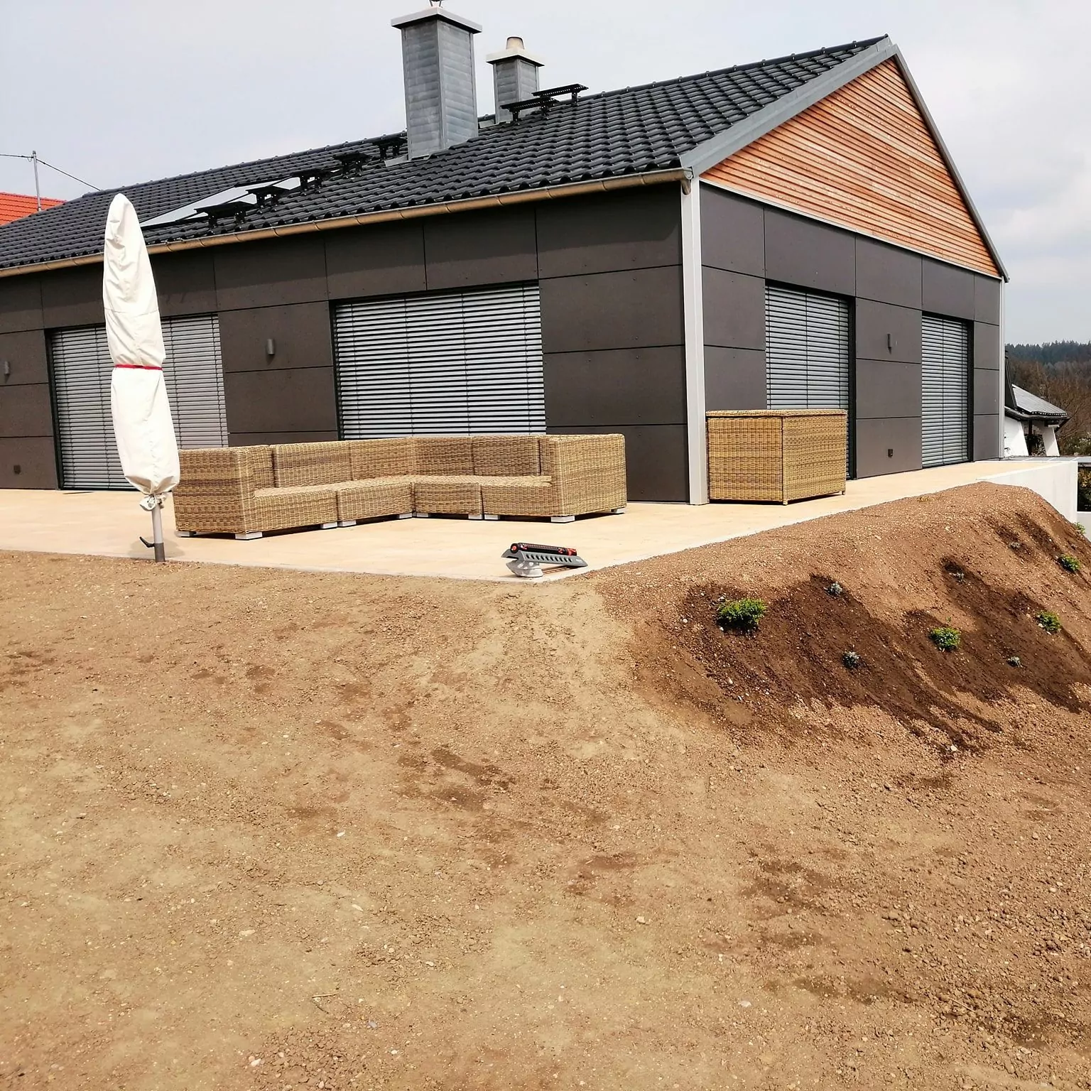 Modernes Haus mit dunkelgrauen Wänden und einem schrägen Dach. Die Terrasse ist mit Korbmöbeln und einem Sonnenschirm ausgestattet. Im Vordergrund ist nackte Erde mit kleinen Pflanzen zu sehen.