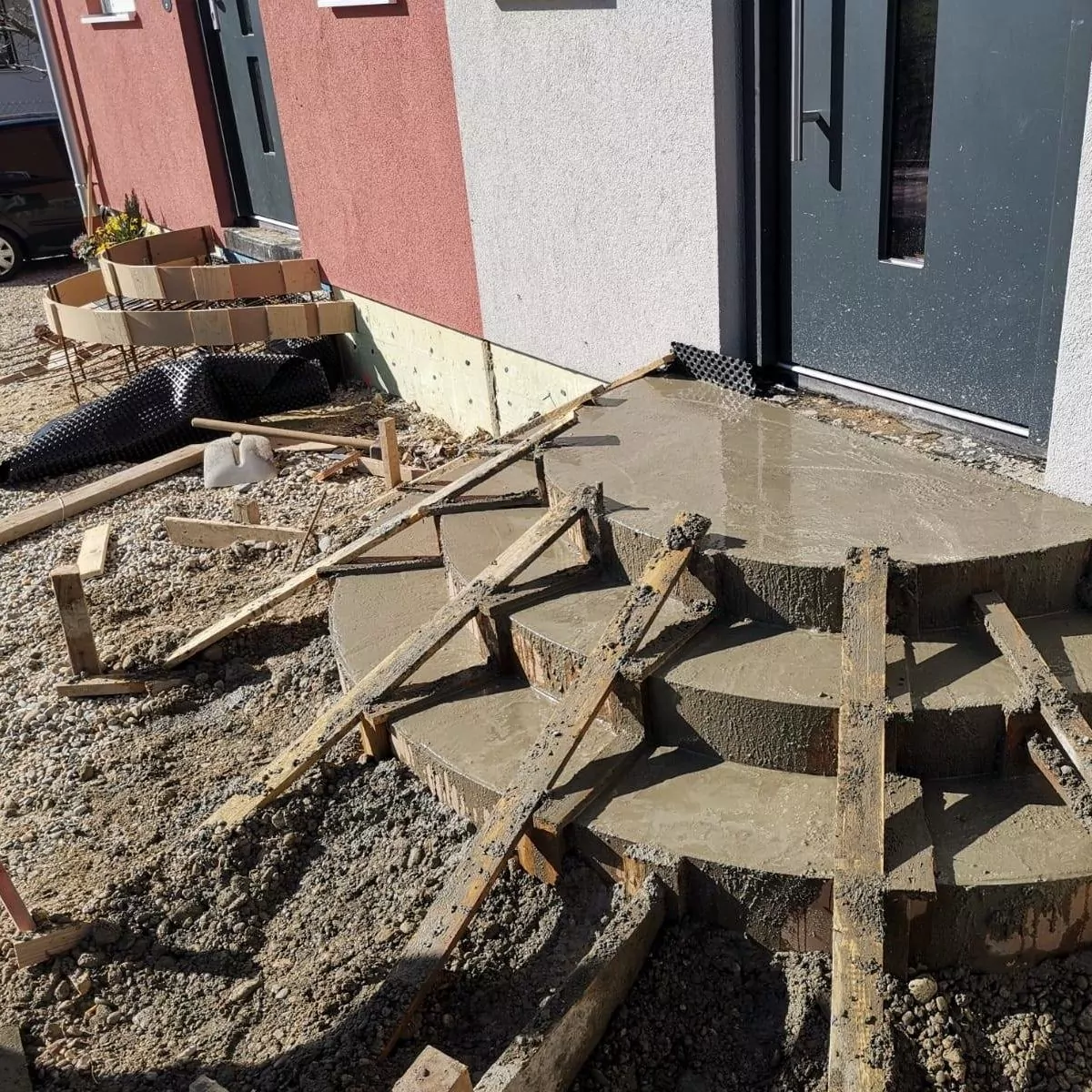 Vor einem Gebäudeeingang werden frische Betonstufen gebaut. Holzformen und Stützen werden eingesetzt, um dem Beton Form zu geben. Im Hintergrund sind Baumaterialien und ein geparktes Auto zu sehen.