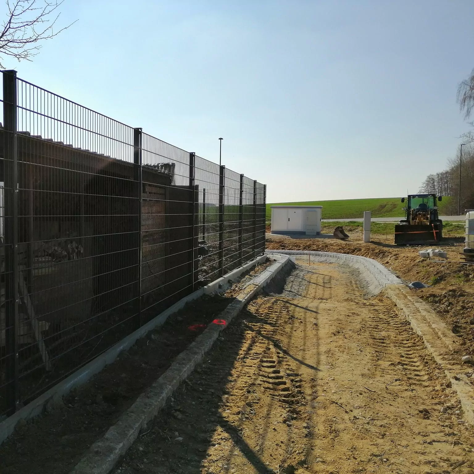 Baustelle mit einer unbefestigten Straße und einem Maschendrahtzaun auf der linken Seite. Im Hintergrund ist neben einem kleinen weißen Gebäude ein Bulldozer zu sehen. Der Himmel ist klar und das Gebiet ist von offenen Feldern umgeben. Auf der unbefestigten Oberfläche sind Spuren zu sehen.