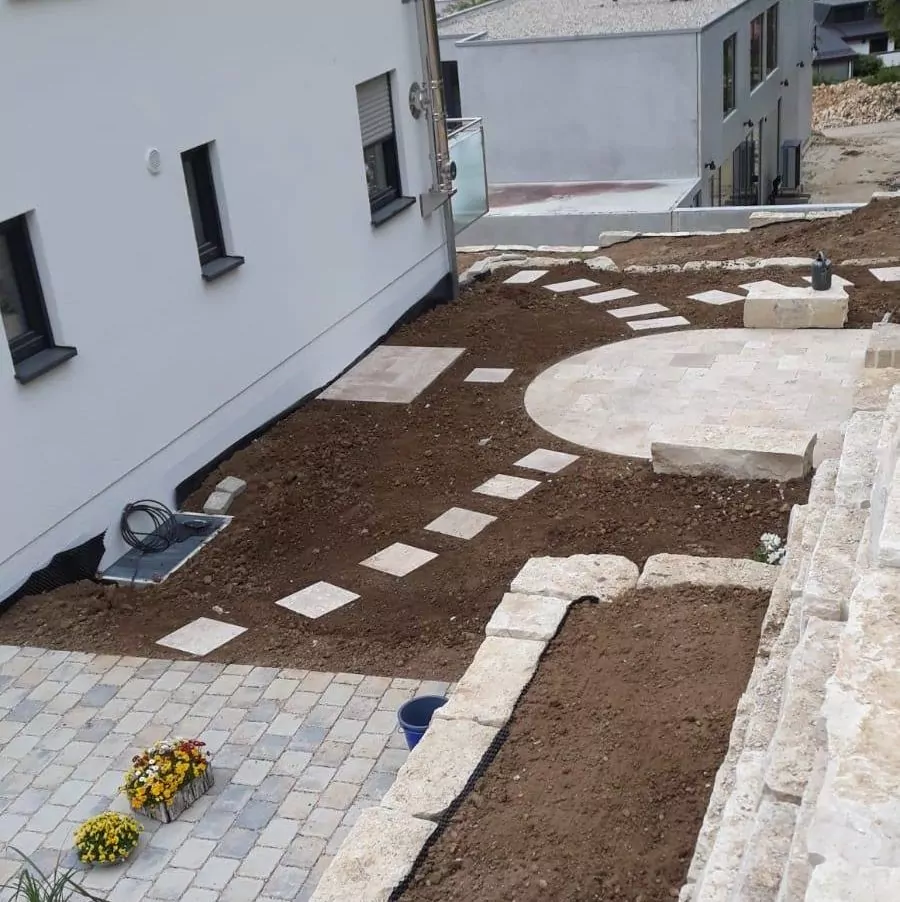 Das Bild zeigt einen neu gestalteten Hinterhof mit einem Feldweg und einer runden Steinterrasse. Auf der rechten Seite befinden sich Steinstufen und in der Ecke ein kleiner Bereich mit blühenden gelben Blumen. Zwei angrenzende Gebäude sind teilweise sichtbar.