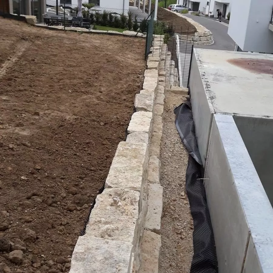 Eine Stützmauer aus großen, rechteckigen Steinen trennt einen unbefestigten Bereich von einer Betonkonstruktion. Schwarzes Schutzgewebe liegt am Ufer, und oben ist ein Maschendrahtzaun zu sehen. Im Hintergrund verläuft eine asphaltierte Straße neben dem Bereich.