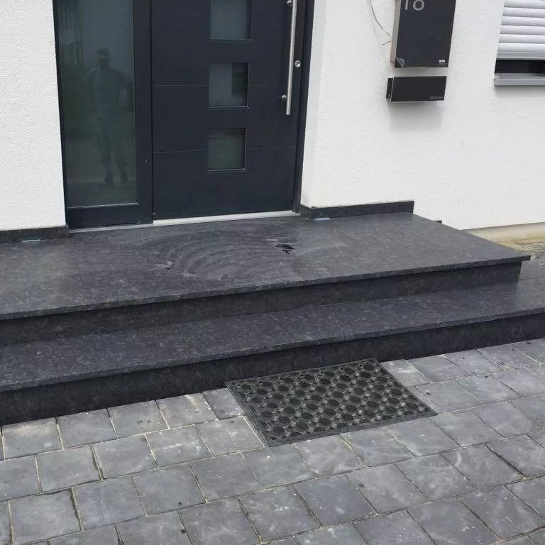 Schwarze Steinstufen führen zu einer modernen schwarzen Tür mit vier quadratischen Fenstern. Auf den Stufen liegt eine Fußmatte, und in der Tür ist schwach das Spiegelbild eines Mannes zu erkennen. Die Stufen sind von grauen Pflastersteinen umgeben.