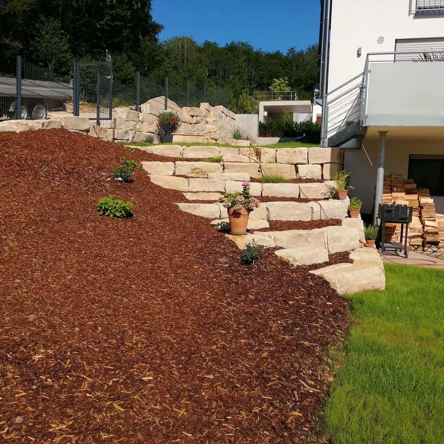 Ein Hanggarten mit großen Steinstufen und Mulch, Topfpflanzen und kleinen Sträuchern. Auf der rechten Seite ist ein weißes Gebäude mit Balkon und am Fuß der Treppe ist ein Stapel Brennholz zu sehen. Der Himmel ist klar und blau.