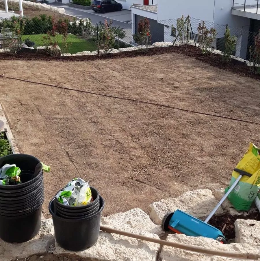 Eine kahle Gartenfläche mit frisch aufbereitetem Boden, umgeben von Steinkanten. Im Vordergrund stehen schwarze Eimer, Säcke mit Pflanzmaterial und ein Gartenschlauch auf dem Boden. Im Hintergrund sind Wohnhäuser und eine Straße zu sehen.