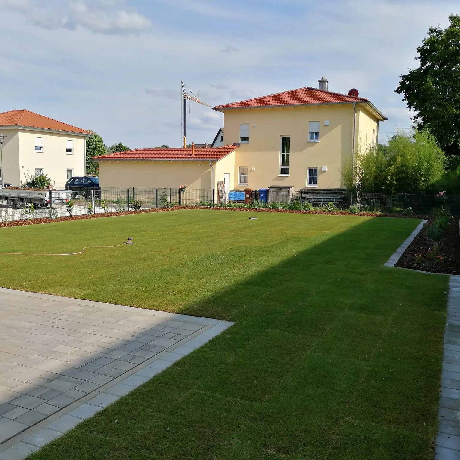 Ein Hinterhof mit einem großen, gepflegten grünen Rasen, der von einem gepflasterten Weg begrenzt wird. Im Hintergrund sind zwei beige Häuser mit roten Dächern zu sehen, und ein Baukran ist zu sehen. Bäume und Sträucher säumen den Rand des Hofes.