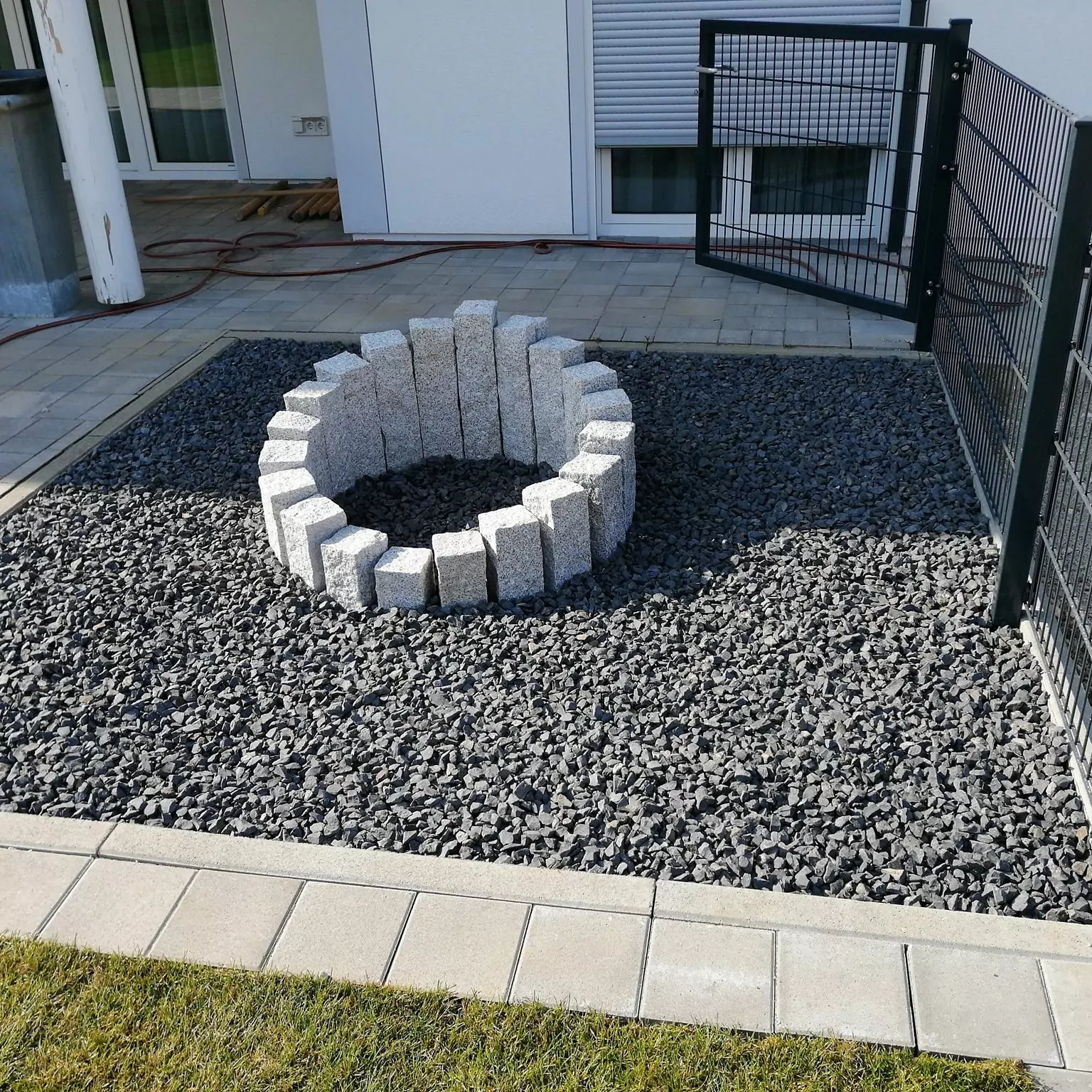 Eine runde Steinstruktur steht auf einer Kiesfläche, umgeben von quadratischen Pflastersteinen. Auf der rechten Seite ist ein Metallzaun mit einem offenen Tor und im Hintergrund ein Gebäude mit weißen Wänden und geschlossenen Fenstern zu sehen.