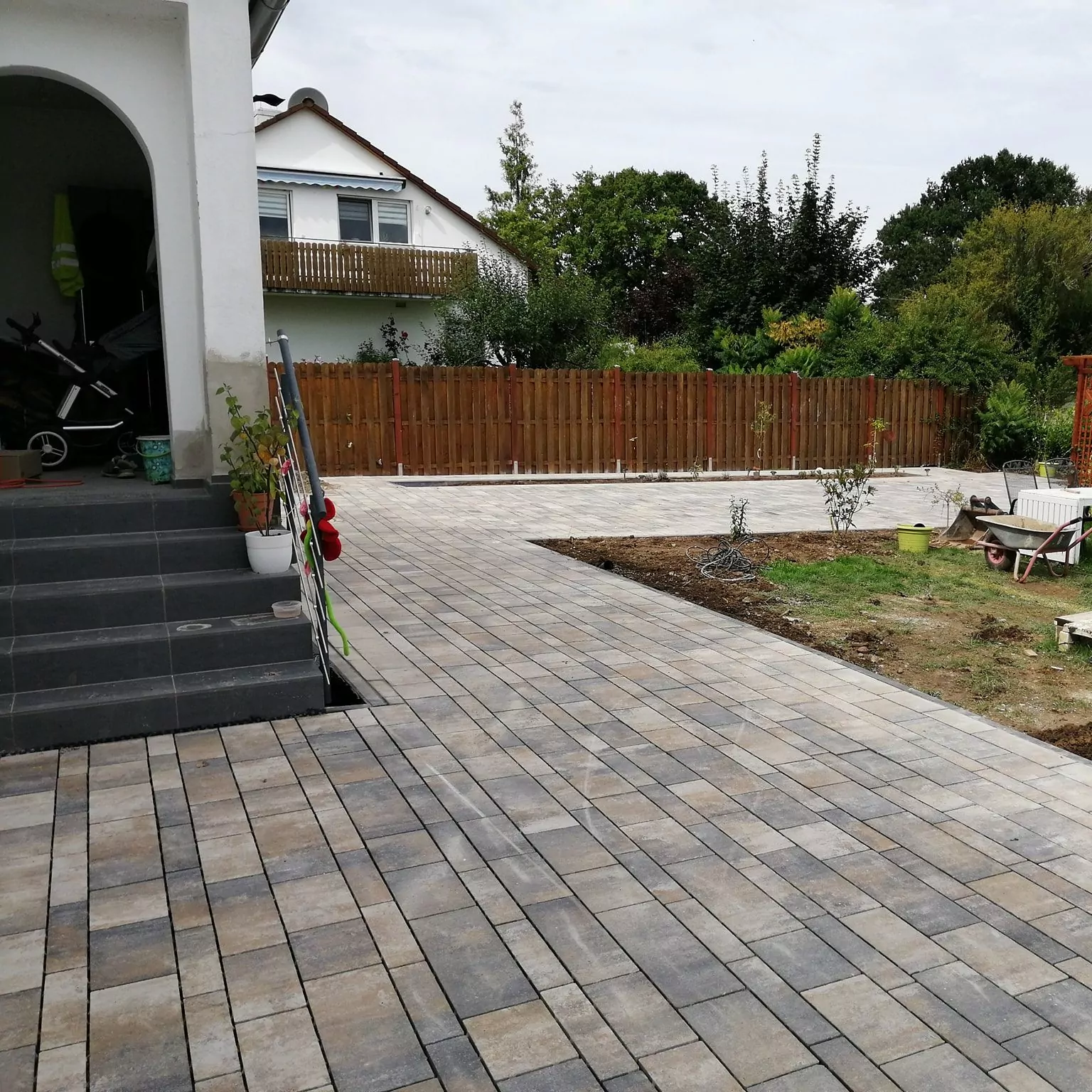 Ein gepflasterter Außenbereich mit grauen Fliesen führt zu einem Holzzaun. Stufen auf der linken Seite führen zu einem gewölbten Eingang eines weißen Gebäudes. Auf der rechten Seite des Bürgersteigs befindet sich ein kleiner Garten mit Gras und einigen Pflanzen. Im Hintergrund ist ein zweistöckiges Haus zu sehen.