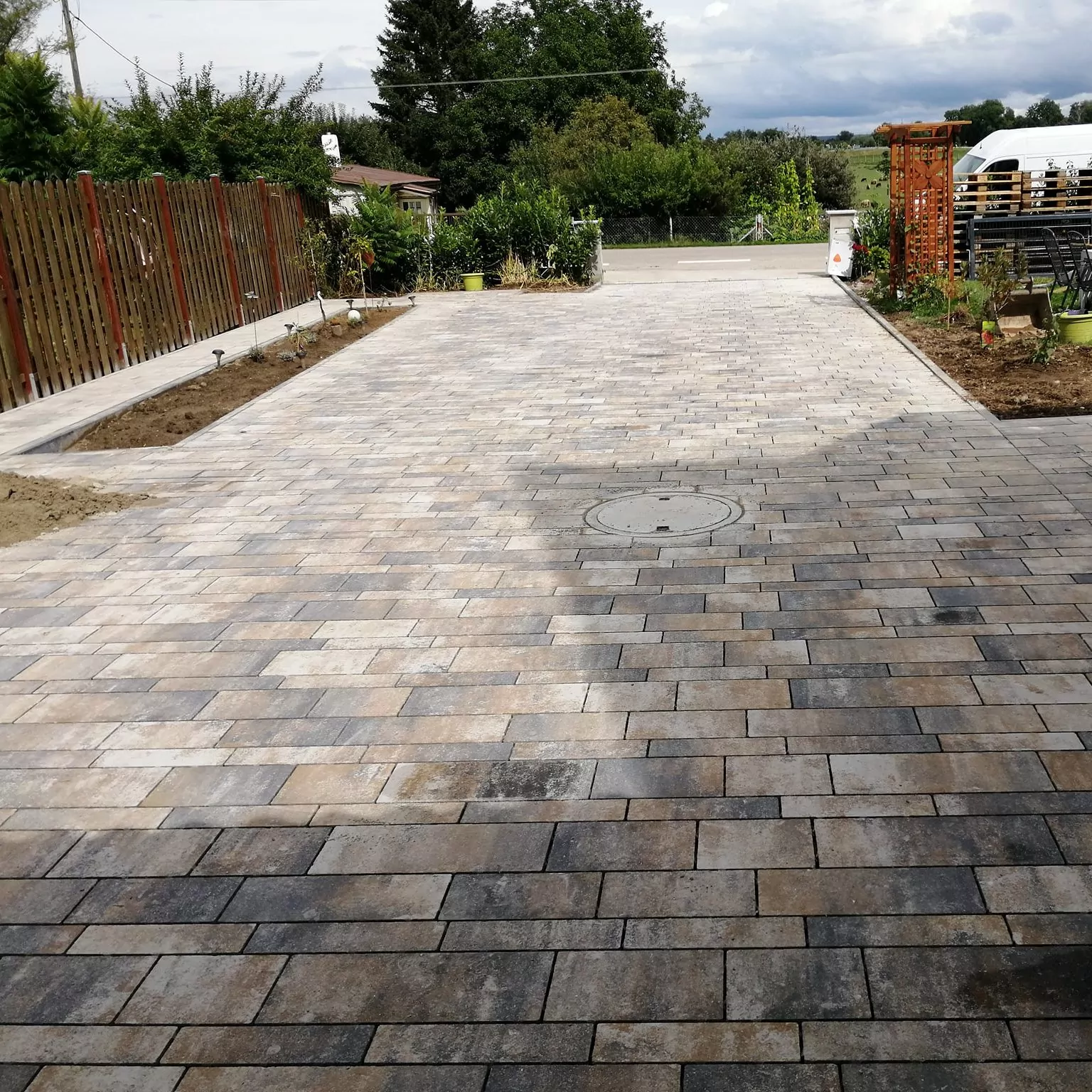 Eine neu gepflasterte Auffahrt aus rechteckigen Steinfliesen in einem Muster aus verschiedenen Grau- und Brauntönen. Die Auffahrt wird auf einer Seite von Gartenbeeten und einem Holzzaun flankiert, im Hintergrund sind Bäume und ein bewölkter Himmel zu sehen.