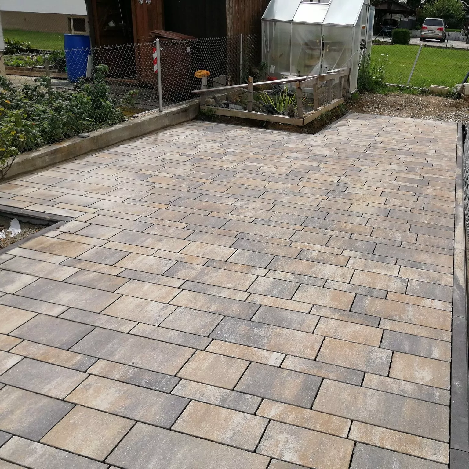 Ein großzügiger Außenbereich mit Verbundsteinpflaster in hellbrauner Farbe. Links ist ein kleiner Garten mit Maschendrahtzaun, im Hintergrund ein Gewächshaus und eine Rasenfläche.