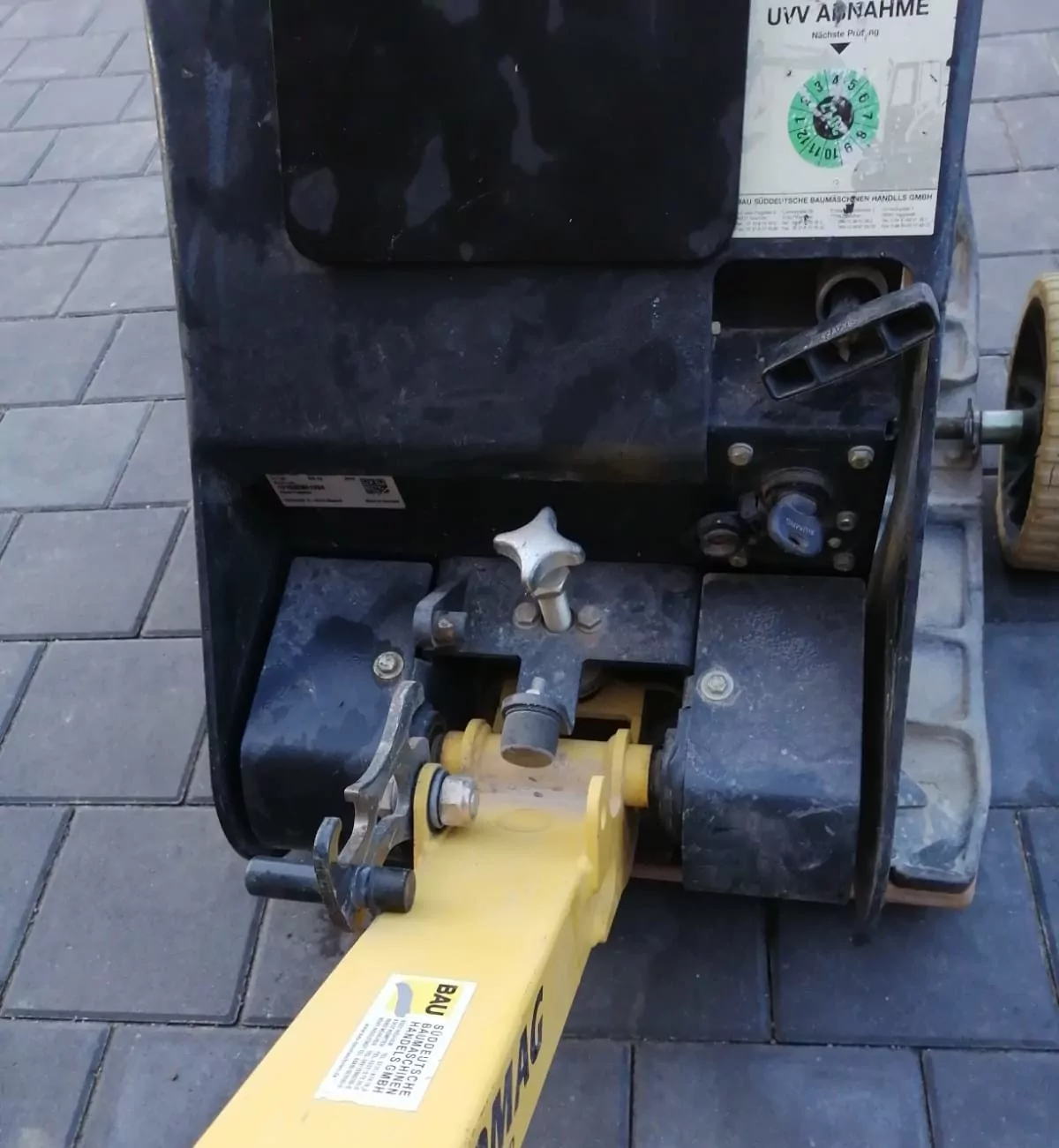 Nahaufnahme eines mechanischen Bauteils auf einer gepflasterten Fläche. Das Gerät hat einen gelben Griff, ein schwarzes Gehäuse, verschiedene Knöpfe und einen Aufkleber mit Text in einer anderen Sprache. Es ist Teil einer Industriemaschine, möglicherweise für den Einsatz im Bauwesen.